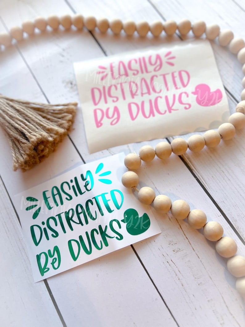 May include: Two vinyl stickers with the text "Easily Distracted by Ducks" in pink and teal lettering. Each sticker features a small duck silhouette.