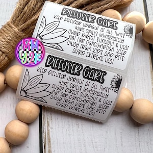 May include: Two white rectangular labels with black text that reads "DIFFUSER CARE". The labels have a floral design and instructions: "Keep diffuser upright at all times, avoid direct sunlight & heat, not for consumption, keep away from kids and pets, avoid extreme heat."
