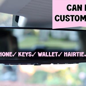May include: A black rearview mirror with a pink sticker that says "PHONE/KEYS/WALLET/HAIR TIE/" and "CAN BE CUSTOMIZED!" in pink text.