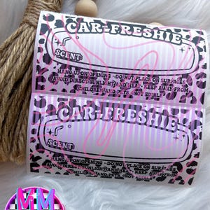May include: Two car air fresheners with the text "CAR-FRESHIE" in a pink and black leopard print design. The background is white with a fluffy texture. The text "SCENT" is also visible.