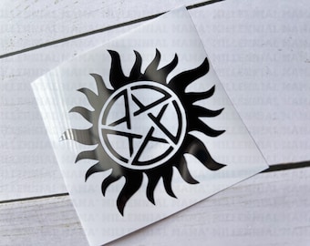 May include: A square adhesive decal featuring a black pentagram symbol enclosed within a circular border, surrounded by stylized black flame-like rays. The design is printed on a clear backing, creating a high-contrast graphic element for application.