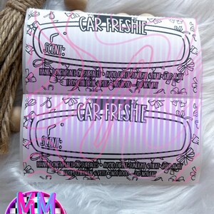 May include: Two car air fresheners with the text "CAR-FRESHIE" in black. The fresheners have a pink and white striped background with pink floral accents. The text "SCENT:" is visible. A decorative tassel and wooden beads are in the background.