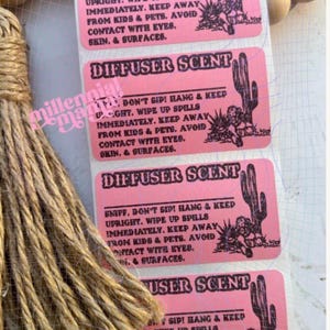May include: Pink rectangular labels with the text "DIFFUSER SCENT" and a cactus illustration. The labels also include safety instructions. A bundle of twine and wooden beads are in the background.