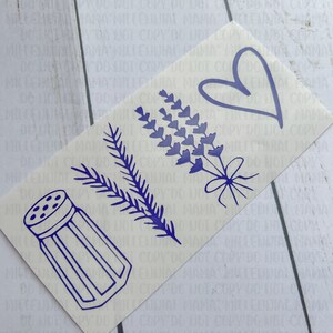 May include: A white sticker with a blue outline of a salt shaker, a sprig of rosemary, a bouquet of lavender, and a heart. The sticker is on a white wooden surface.