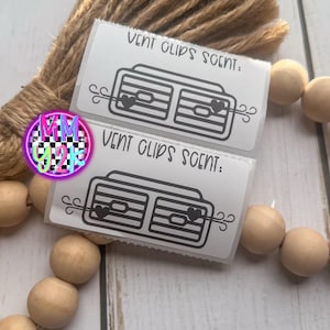 May include: Two white rectangular vent clip scent packages with black line art of a car vent and heart. The text "VENT CLIPS SCENT:" is printed on each package. The packages are displayed with wooden beads and a brown rope.
