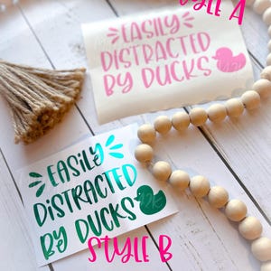 May include: Two versions of a vinyl sticker with the text "Easily Distracted by Ducks" and a rubber duck graphic. The top sticker is pink and the bottom sticker is teal.