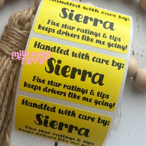 May include: A roll of yellow stickers with the name "Sierra" in large black letters. The stickers also include the text "Handled with care by:" and "Five star ratings & tips keeps drivers like me going!" Wooden beads and twine are in the background.