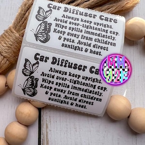 May include: Two white labels with black text and butterfly illustrations, reading "Car Diffuser Care." Instructions include keeping upright, avoiding over-tightening, wiping spills, and avoiding sunlight and heat. The labels are next to a bundle of twine and wooden beads.
