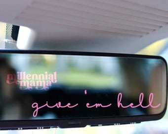 May include: A rectangular vehicle rearview mirror featuring pink vinyl lettering. The text reads millennial mama in a bold, rounded font and give 'em hell in a cursive script. The mirror is mounted beneath a light gray vehicle ceiling panel.