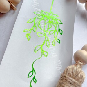 May include: A neon green plant decal on a white background. The decal features a detailed outline of a hanging plant with leaves and a central flower-like structure. The text "Mierma Yok" is visible in the background.