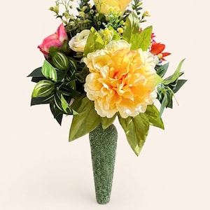 May include: A floral arrangement featuring a large, central yellow peony, smaller yellow roses, and pink blossoms. The bouquet includes various green leaves and stems, all secured within a textured, cone-shaped green base with a granular surface.
