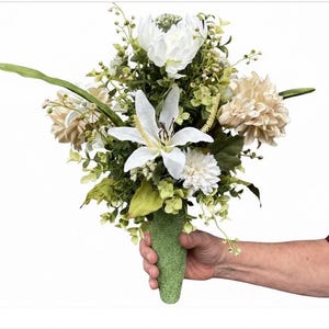 May include: A floral arrangement featuring white lilies, cream-colored dahlias, and various green foliage. The flowers are gathered into a textured, light green, cone-shaped base. The bouquet includes small white buds and leafy stems.