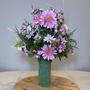 May include: A floral arrangement featuring pink and white daisies, small purple flowers, and green foliage. The flowers are arranged in a green vase with a speckled texture. The arrangement is set on a wooden surface.