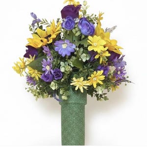 May include: A floral arrangement featuring a mix of yellow sunflowers, purple roses, and smaller blue and yellow daisies with green foliage. The flowers are set into a cylindrical, glittery green vase with a textured surface and a flared top rim.