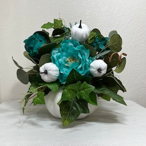 May include: A white pumpkin-shaped vase filled with teal and green artificial flowers and white pumpkins. The arrangement includes large teal peonies, smaller teal flowers, and green foliage, creating a fall-themed centerpiece.