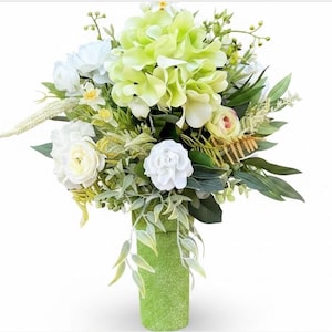 May include: A floral arrangement featuring large light green hydrangea blooms, white roses, and various green foliage. The bouquet is displayed in a cylindrical, textured light green vase with a glittery finish, set against a plain white background.