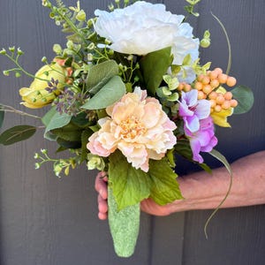 May include: A floral bouquet featuring a large white peony, a soft peach peony, yellow blossoms, purple flowers, and clusters of small orange berries. The arrangement includes various green leaves and stems, all gathered into a textured green handle.