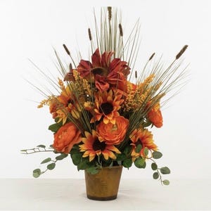 May include: A floral arrangement in a brown pot featuring orange and red sunflowers, round orange ranunculus flowers, yellow filler sprigs, green eucalyptus leaves, and tall, thin, upright blades of grass extending from the center.