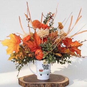 May include: A white ceramic mug featuring the text Hello Fall and a graphic of a light blue truck carrying a pumpkin. The mug is filled with an arrangement of artificial autumn foliage, including orange maple leaves, berries, and small pumpkins.