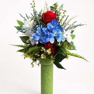 May include: A vibrant floral arrangement featuring a red rose, blue hydrangeas, and white flowers, accented with green foliage. The bouquet is presented in a textured green vase, creating a striking contrast against the white background.