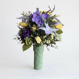 May include: A bouquet of artificial flowers featuring purple hydrangeas, yellow roses, lavender sprigs, and green foliage arranged in a tall, cylindrical, speckled light green vase with a textured, matte finish and a flared top rim.