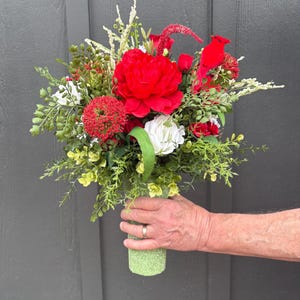 May include: A vibrant bouquet featuring a large red peony as the focal point, complemented by white and red accent flowers, and lush green foliage. The bouquet is held in a light green, textured vase, creating a striking contrast against the dark background.
