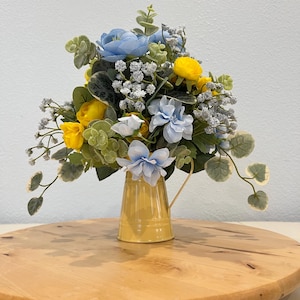 May include: A yellow metal pitcher filled with a floral arrangement of blue and yellow flowers. The flowers are arranged in a cluster and include baby's breath. The pitcher is sitting on a wooden surface.