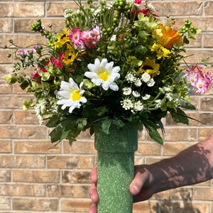 May include: A vibrant floral arrangement featuring a variety of colorful flowers, including daisies, sunflowers, and pink blooms, atop a textured green vase. The vase is held by a person's hand. The flowers are a mix of yellow, white, and pink.