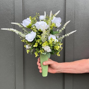 May include: A bouquet of white roses and greenery, held in a light green, textured cone-shaped holder. The arrangement includes white flowers, small white buds, and various shades of green foliage. The background is a gray wooden door.