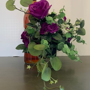May include: A decorative arrangement of purple roses, small purple flowers, and green foliage. The floral display is placed in front of a wine bottle. The bottle has a red cap and a clear glass body. The arrangement is on a dark brown surface.