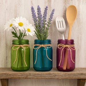 May include: Three colored mason jars on a wooden shelf. The jars are green, teal, and purple, each tied with twine. The green jar holds white daisies, the teal jar has lavender, and the purple jar contains a wooden spoon and a slotted metal spatula.