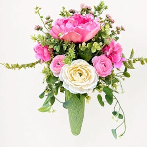 May include: A floral arrangement featuring a large bright pink peony, a central cream-colored ranunculus, and smaller pink ranunculus blossoms. The bouquet includes green foliage, small sprigs of greenery, and is held in a textured green cone.