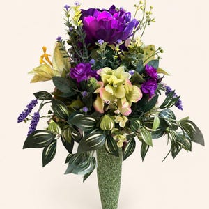 May include: A floral arrangement featuring a large deep purple peony, pale yellow hydrangeas, and small purple blossoms. The bouquet includes green leaves with white stripes and is displayed in a textured, light green, tapered vase.