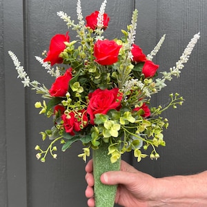 May include: A bouquet of red roses and white flowers with green foliage. The bouquet is held in a green, textured, cylindrical holder. The flowers are vibrant against the dark background.