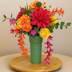 May include: A vibrant floral arrangement in a green vase. The bouquet features a mix of orange, yellow, pink, and red flowers, including roses, dahlias, and geraniums. The flowers are arranged in a vase with a textured green finish.