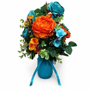 May include: A vibrant floral arrangement in a turquoise mason jar. The bouquet features large orange roses, teal and light blue roses, and smaller yellow and orange flowers, with green foliage. A turquoise ribbon is tied around the jar.