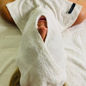 May include: A person's face is covered with a white towel, with only the nose and mouth visible. The towel is soft and fluffy, suggesting a spa or skincare treatment. The person is lying on a white surface, possibly a massage table.
