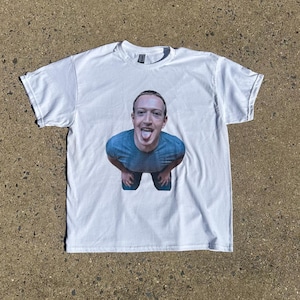 May include: White t-shirt with a full-colour image of a person with their tongue out. The person is wearing a blue shirt and dark trousers. The t-shirt is lying flat on a concrete surface.