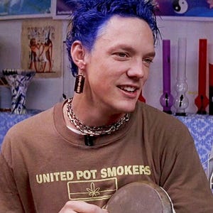 May include: A person with blue hair wearing a brown t-shirt with the text "UNITED POT SMOKERS" and a cannabis leaf graphic. They are wearing a silver chain necklace and earrings. Various glass pipes are visible in the background.