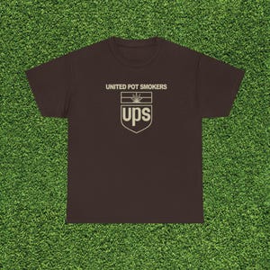 May include: A brown t-shirt with the text "UNITED POT SMOKERS" above a logo that resembles the UPS logo, but with a cannabis leaf and the letters "ups". The shirt is displayed on a green surface.