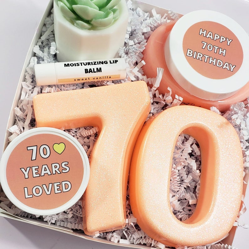 May include: A gift box filled with items for a 70th birthday celebration. The box contains two large peach-colored numbers 7 and 0, a small jar with a peach-colored lid and a label that says "70 Years Loved", a small jar with a white lid and a label that says "Happy 70th Birthday", a small succulent plant in a white pot, and a small tube of lip balm with a label that says "Moisturizing Lip Balm Sweet Vanilla".