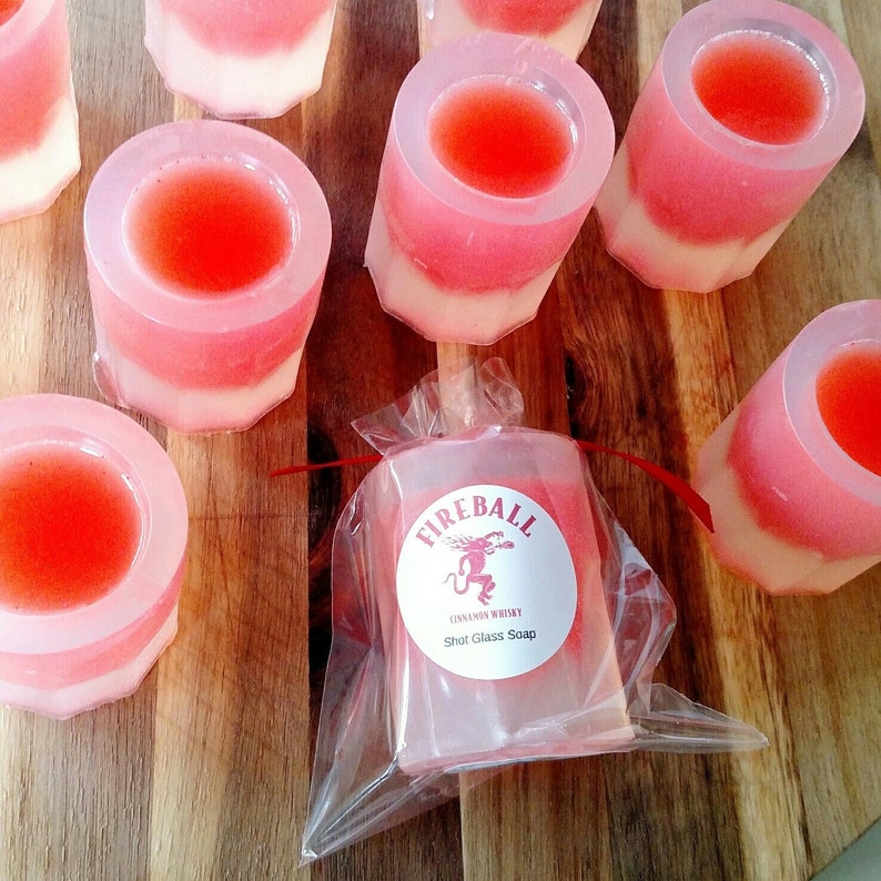 May include: Shot glass soaps in a variety of colors, including red, pink, and white, are arranged on a wooden surface. One soap is packaged in clear plastic with a label that reads "Fireball Cinnamon Whisky Shot Glass Soap."