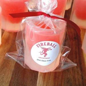 May include: A shot glass-shaped soap, packaged in clear cellophane and tied with a red ribbon. The soap is a gradient of pink and white, with a circular label that reads "Fireball Cinnamon Whisky Shot Glass Soap" and features a red dragon.