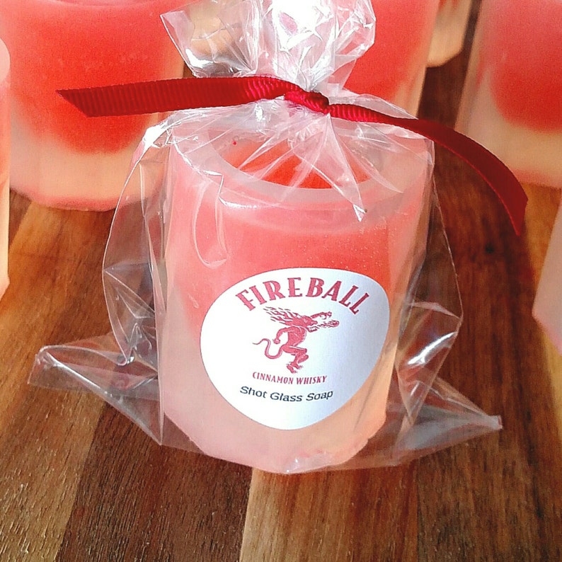 May include: A shot glass-shaped soap, packaged in clear cellophane and tied with a red ribbon. The soap is a gradient of pink and white, with a circular label that reads "Fireball Cinnamon Whisky Shot Glass Soap" and features a red dragon.