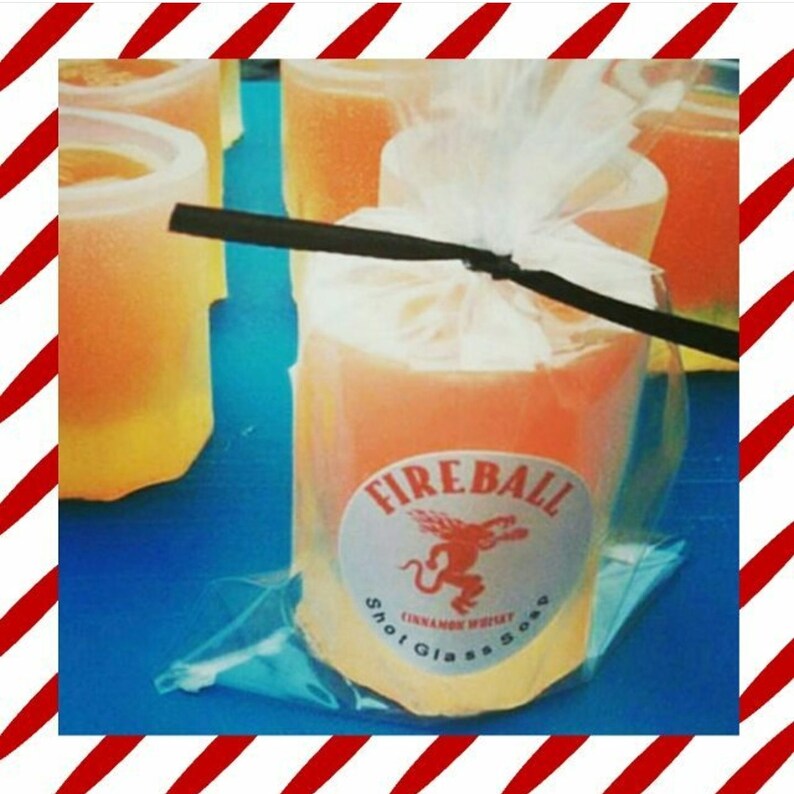 May include: Orange-colored shot glass soaps, packaged in clear cellophane, with a white label that reads "Fireball Cinnamon Whiskey Shot Glass Soap". The label features a red dragon logo. The soaps are on a blue surface.