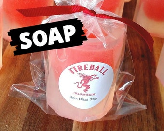 May include: A red and white shot glass shaped soap bar with a Fireball Cinnamon Whisky logo. The soap is wrapped in clear plastic and tied with a red ribbon. The text on the soap reads "Fireball Cinnamon Whisky Shot Glass Soap".