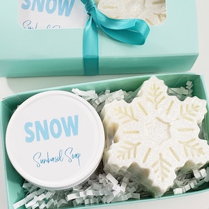 May include: A gift box with a snowflake-shaped soap and a jar of Sunbasil Soap. The box is light blue and tied with a teal ribbon. The soap is white and the jar has the word "SNOW" in light blue.