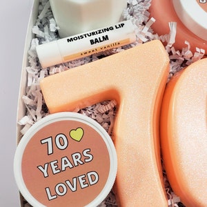 May include: A gift box filled with peach-colored bath products, including two large bath bombs shaped like the numbers 7 and 0, a small jar of body butter with the text "70 Years Loved" and a lip balm labeled "Moisturizing Lip Balm, Sweet Vanilla".