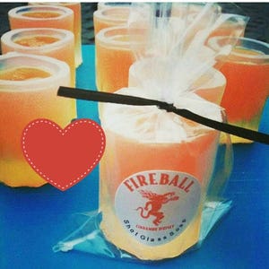 May include: Orange-colored shot glass soaps, some wrapped in clear cellophane. The label reads "Fireball Cinnamon Whiskey Shot Glass Soap" with a red dragon logo. The soaps are arranged on a blue surface.