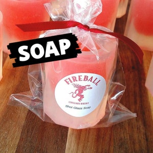 May include: A shot glass-shaped soap, packaged in clear cellophane and tied with a red ribbon. The soap is a gradient of pink and white, with a label that reads "Fireball Cinnamon Whisky Shot Glass Soap."